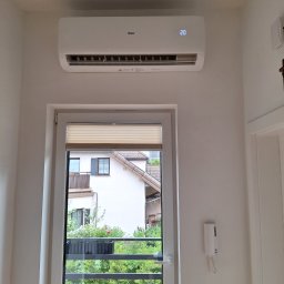 SUN ENERGY ŁUKASZ KACZMAREK - Biały klimatyzator Haier zamontowany nad oknem balkonowym z widokiem na budynek mieszkalny i zieleń, na wyświetlaczu widoczna temperatura 20 stopni Celsjusza.