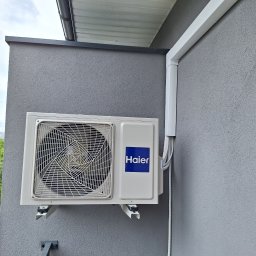 SUN ENERGY ŁUKASZ KACZMAREK - Zewnętrzna jednostka klimatyzacji Haier zamontowana na szarej, teksturowanej ścianie budynku, widoczne białe rury instalacyjne i fragment zieleni w tle.