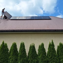 SUN ENERGY ŁUKASZ KACZMAREK - Instalator na dachu z brązowej blachy trapezowej montuje panel fotowoltaiczny; widoczna drabina oparta o dach i rząd zielonych tuj na tle beżowej ściany budynku.