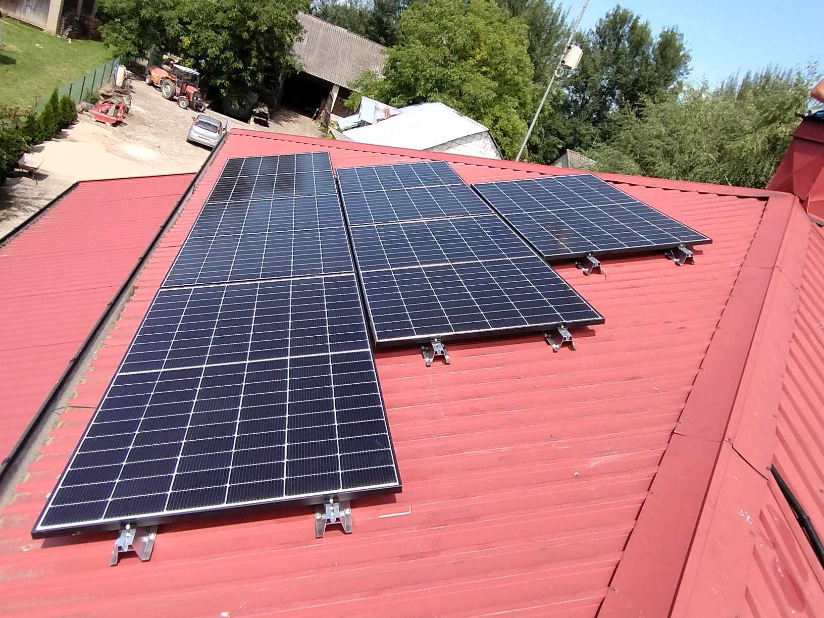 Panele fotowoltaiczne zamontowane na czerwonym, blaszanym dachu budynku gospodarczego na wsi, widoczne wsporniki mocujące.