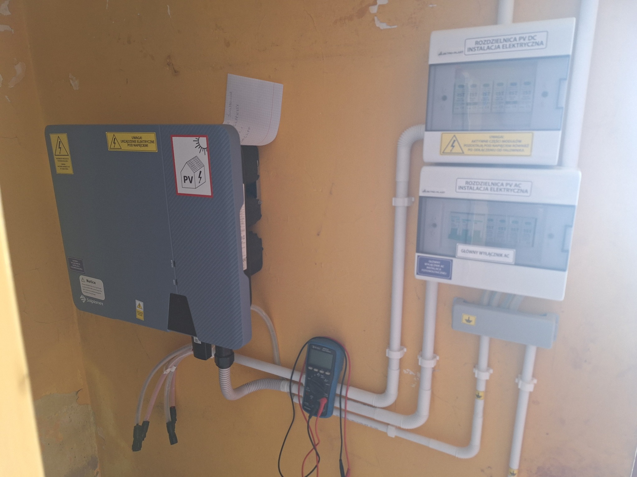 Szary inwerter fotowoltaiczny Solplanet zamontowany na pomarańczowej ścianie obok rozdzielnic PV DC i AC, z podłączonym multimetrem cyfrowym do pomiaru napięcia.