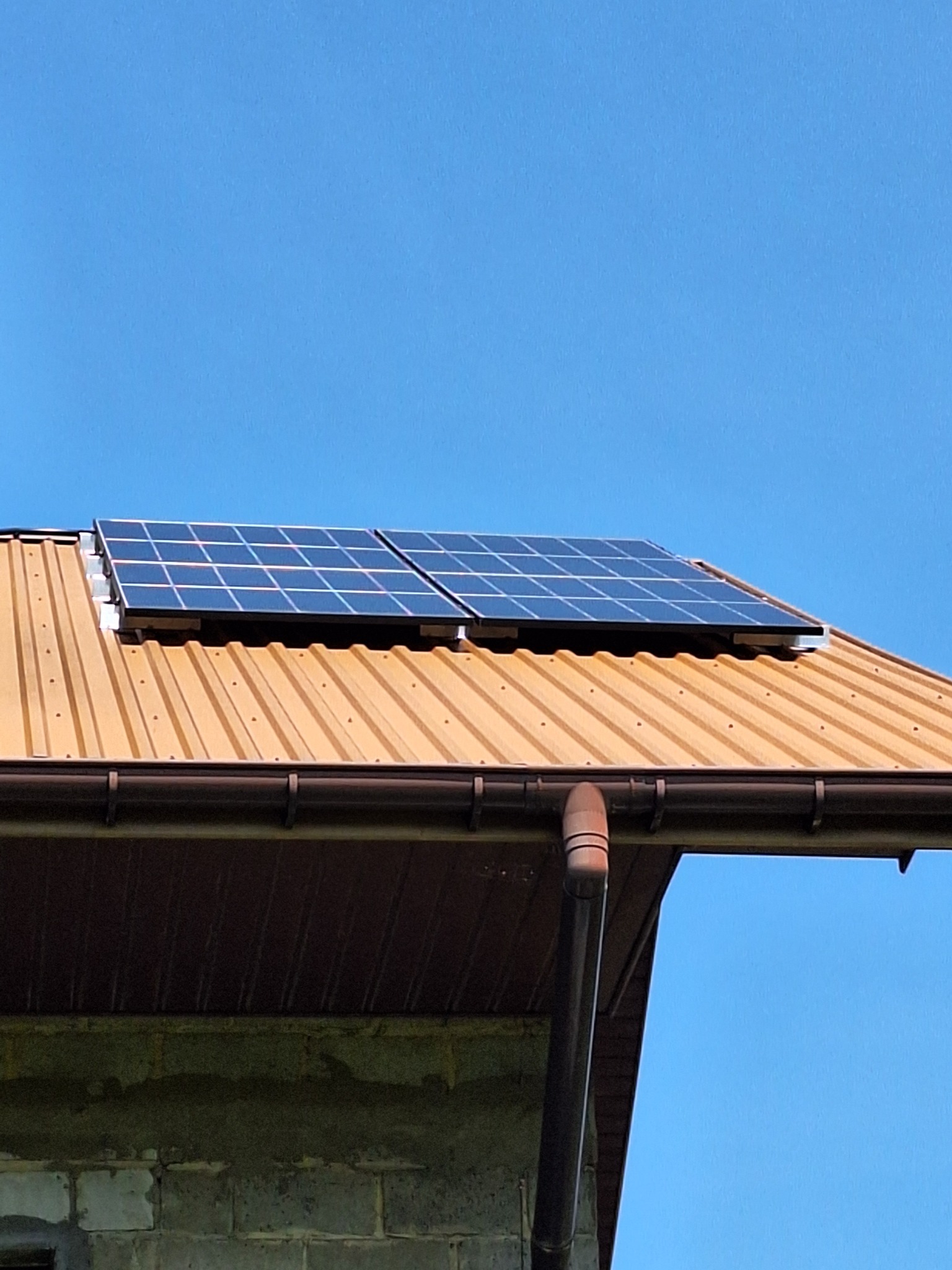 Dwa panele fotowoltaiczne zamontowane na brązowym, metalowym dachu budynku z widoczną rynną spustową, na tle błękitnego nieba.