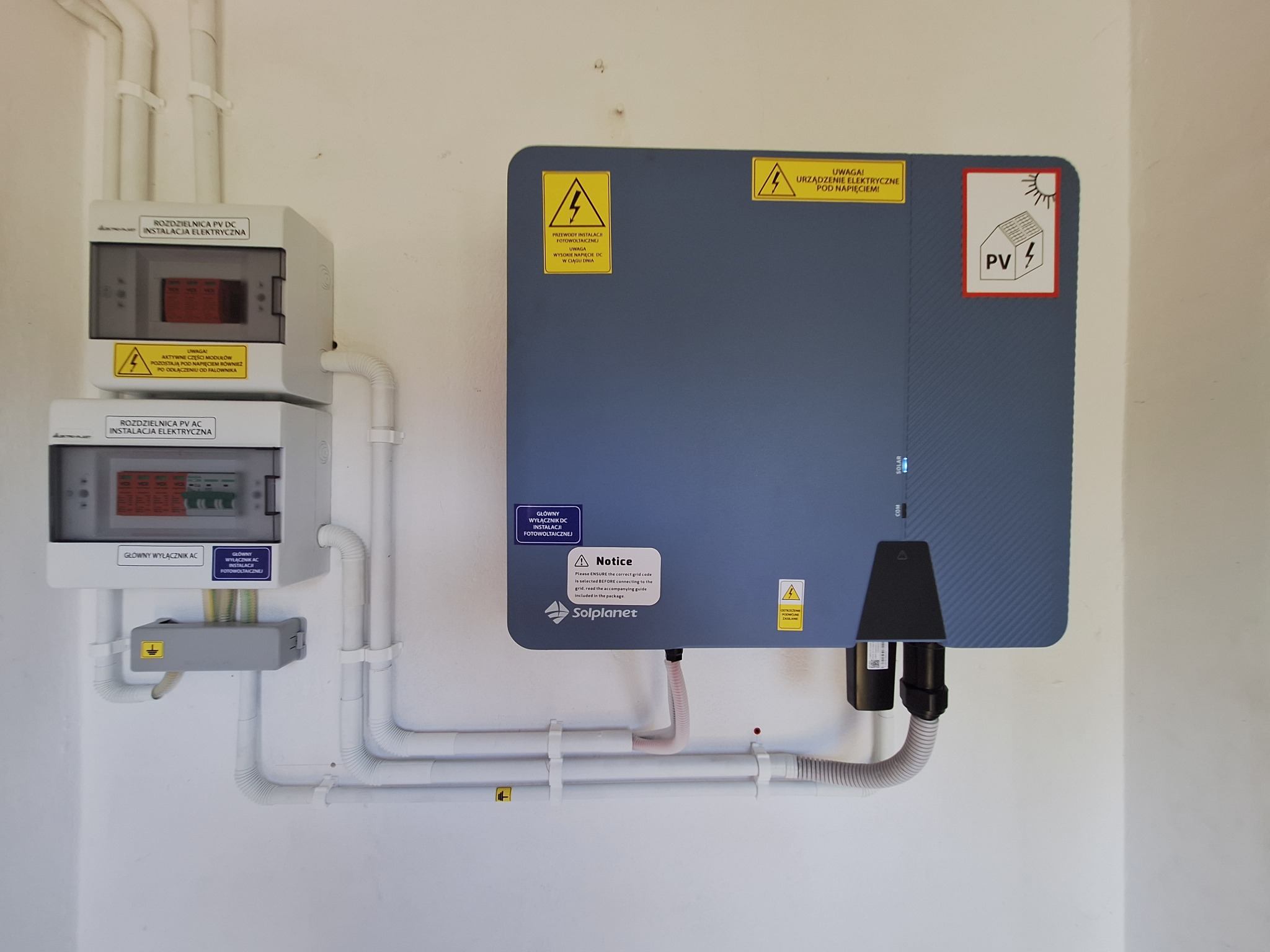 Rozdzielnice PV DC i AC, falownik Solplanet z ostrzeżeniami o napięciu, instalacja elektryczna wewnątrz budynku, białe rury osłonowe.