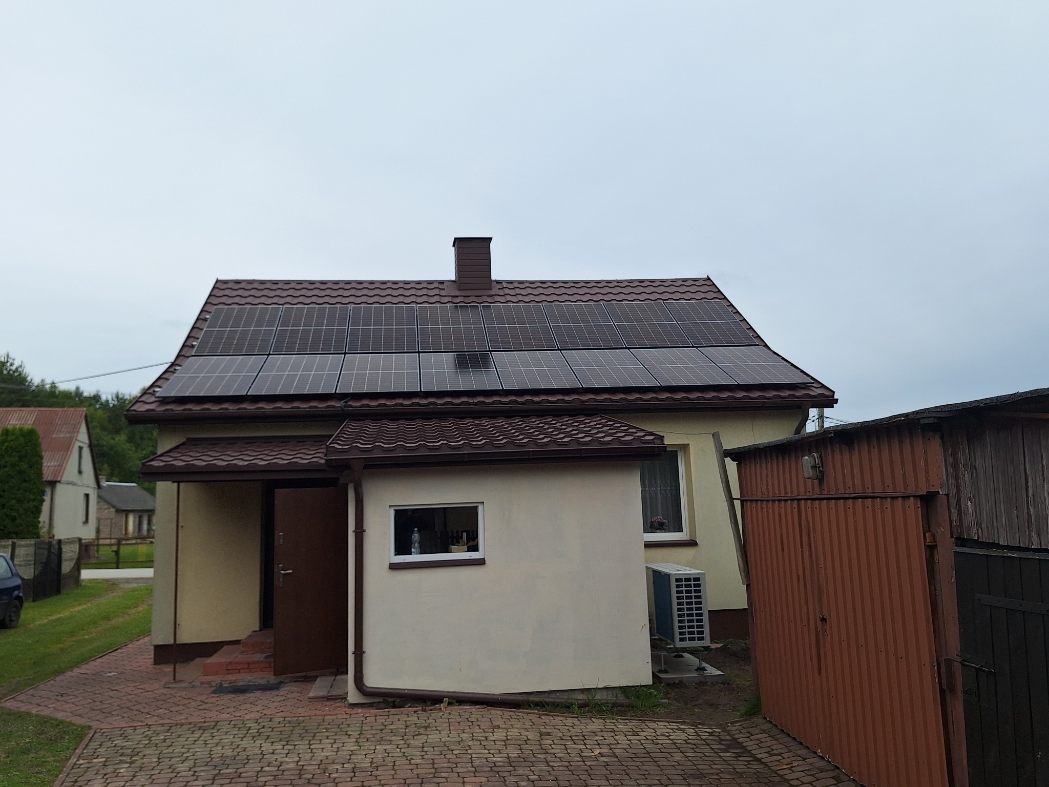 Dom jednorodzinny z brązowym dachem pokrytym panelami fotowoltaicznymi, widoczny komin i jednostka zewnętrzna klimatyzacji, obok garaż blaszany.