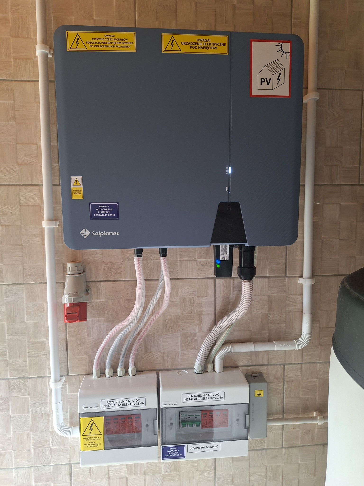 Szczegółowe ujęcie instalacji fotowoltaicznej w Luta, prezentujące falownik marki Solplanet, rozdzielnice PV DC i AC, oraz okablowanie na tle płytek imitujących drewno.