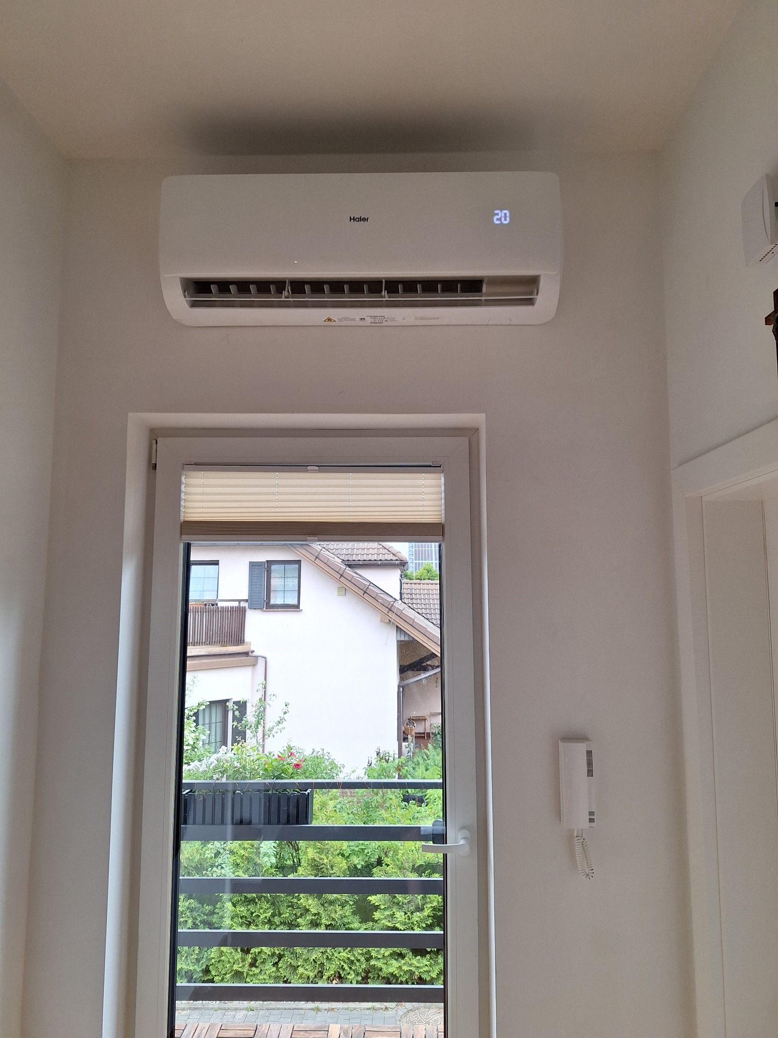 Biały klimatyzator Haier zamontowany nad oknem balkonowym z widokiem na budynek mieszkalny i zieleń, na wyświetlaczu widoczna temperatura 20 stopni Celsjusza.