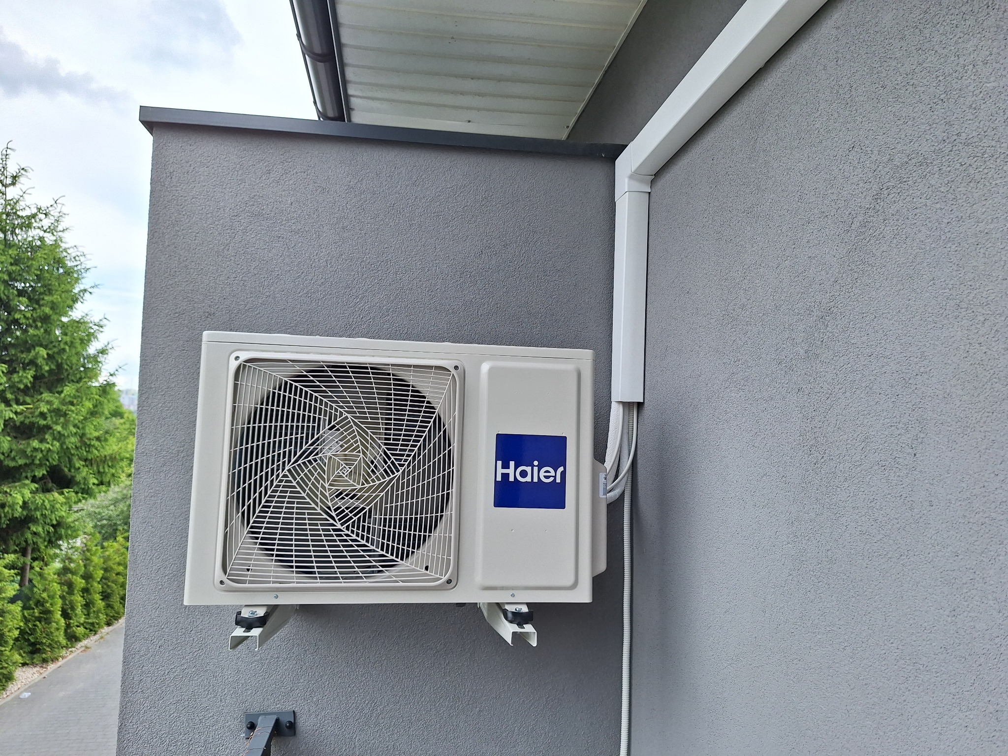 Zewnętrzna jednostka klimatyzacji Haier zamontowana na szarej, teksturowanej ścianie budynku, widoczne białe rury instalacyjne i fragment zieleni w tle.