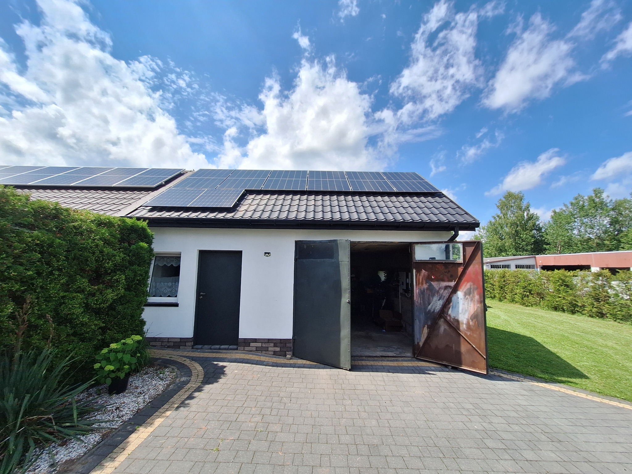Dom jednorodzinny z panelami słonecznymi na dachu, widok z chodnika, otwarte wrota garażowe po prawej stronie.