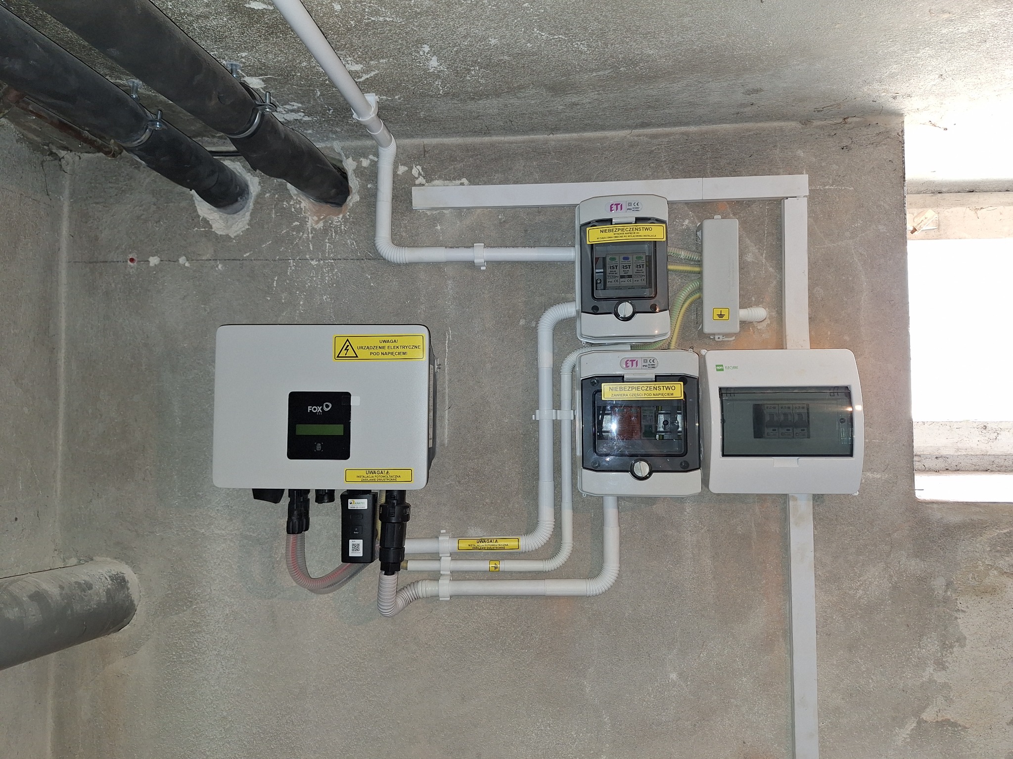 Instalacja fotowoltaiczna: falownik FOX ESS, zabezpieczenia ETI i skrzynka rozdzielcza na surowej ścianie z widocznymi rurami instalacyjnymi
