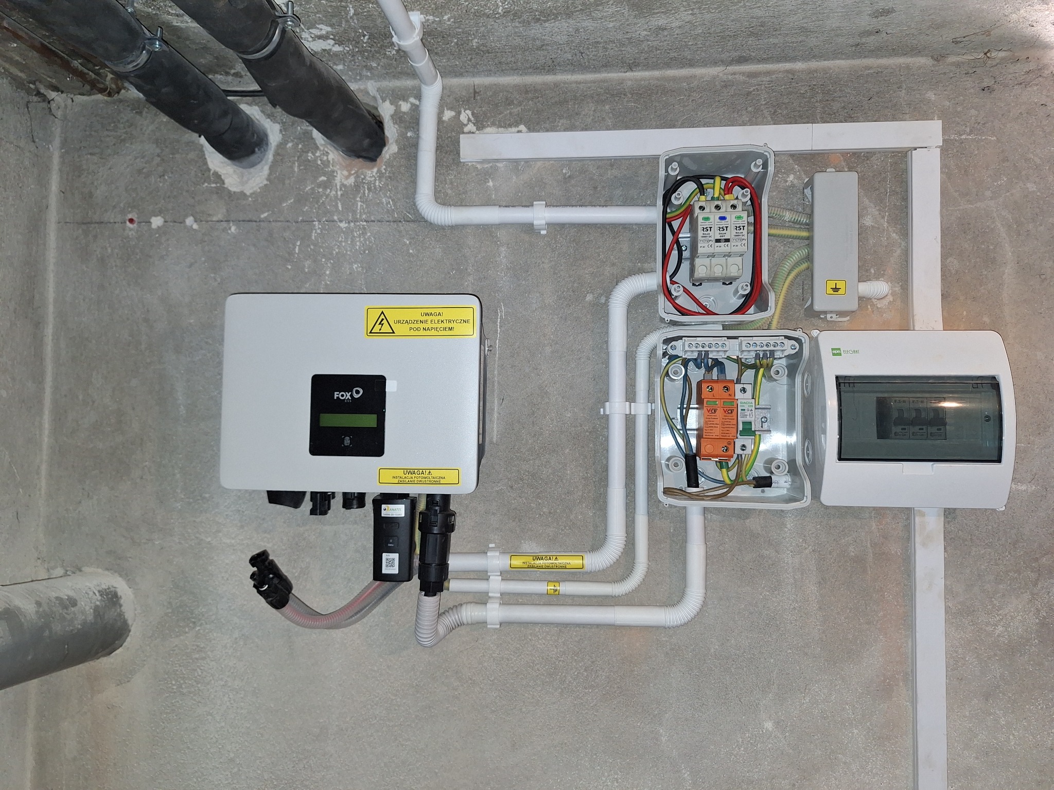 Szary inwerter marki FOXO z żółtą naklejką ostrzegawczą, podłączony przewodami do rozdzielni elektrycznej z zabezpieczeniami, zamontowany na surowej, betonowej ścianie wewnątrz budynku. Widoczne...