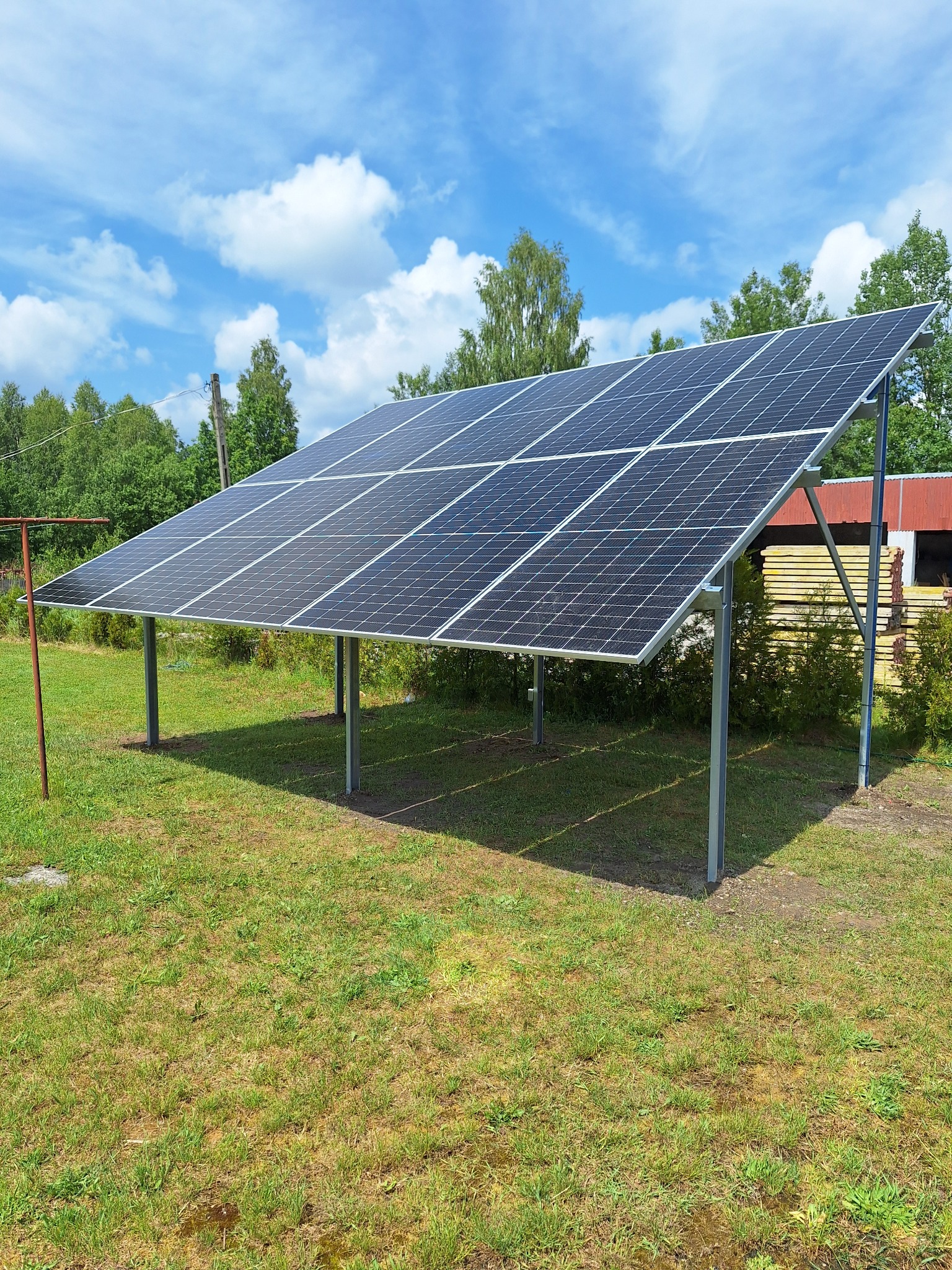 Panele fotowoltaiczne zamontowane na metalowej konstrukcji na trawiastym terenie, błękitne niebo z chmurami w tle.