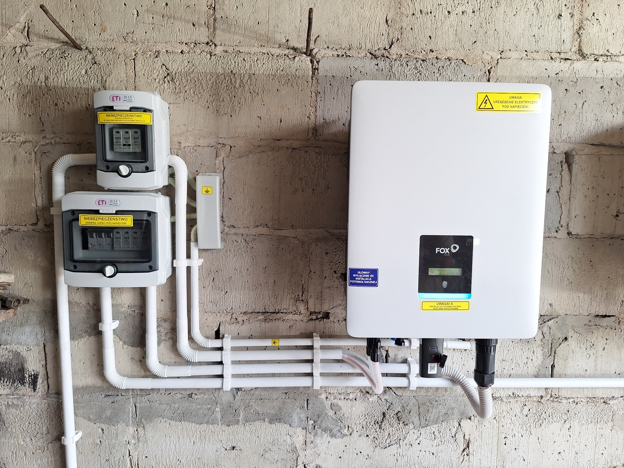 Zamontowany inwerter fotowoltaiczny marki Fox ESS oraz rozdzielnice elektryczne ETI na surowej, betonowej ścianie. Widoczne rury instalacyjne i żółte naklejki ostrzegawcze.