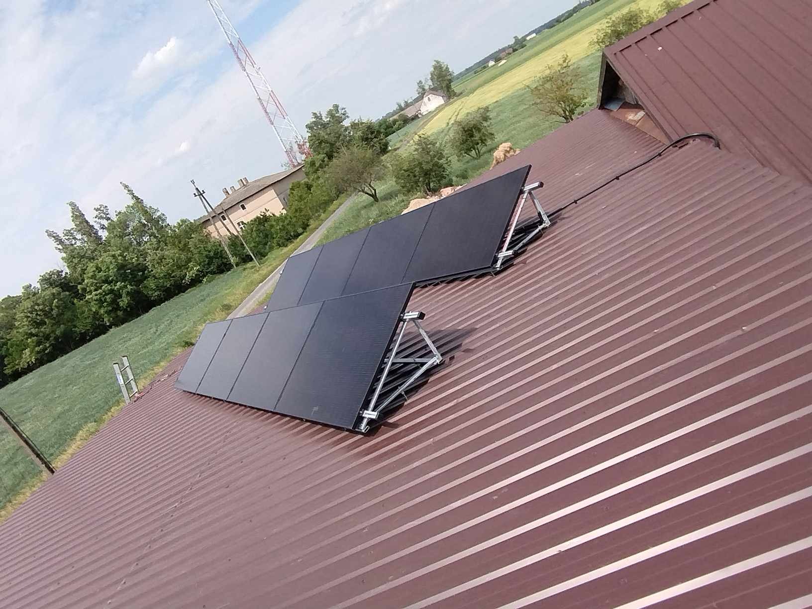 Panele fotowoltaiczne zamontowane na brązowym, blaszanym dachu budynku gospodarczego; w tle zielone pola i maszt telekomunikacyjny.