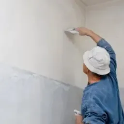 Mężczyzna w niebieskiej bluzie i białym kapeluszu nakłada szpachlą gładź na ścianę w jasnym pomieszczeniu, widoczny etap pracy z częściowo wykończoną powierzchnią.
