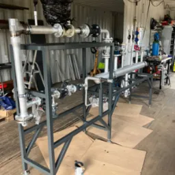 Konstrukcja metalowa z rurami i zaworami, prawdopodobnie element instalacji przemysłowej, w trakcie montażu lub testowania w warsztacie, z widocznymi narzędziami i materiałami w tle.