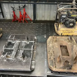 Warsztatowe ujęcie nowej, błyszczącej, spawanej płyty wibracyjnej obok starej, zardzewiałej płyty na perforowanym stole spawalniczym, z podnośnikami samochodowymi w tle.