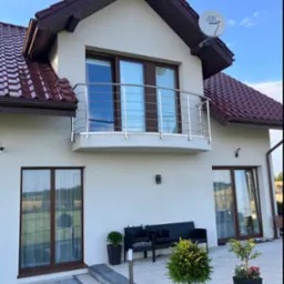 Nowoczesny dom z balkonem z balustradą ze stali nierdzewnej, brązowymi oknami i dachem z dachówki, widok z poziomu tarasu z meblami ogrodowymi i roślinami w donicach.