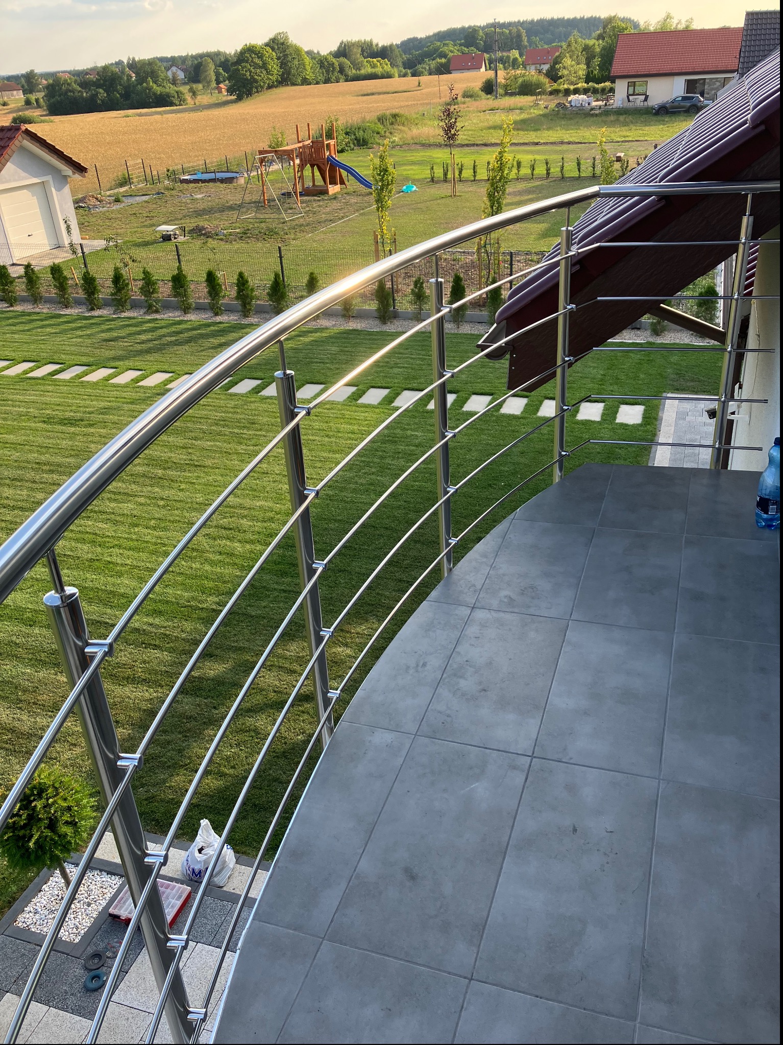 Nowoczesna, zakrzywiona balustrada ze stali nierdzewnej na tarasie z szarych płytek, z widokiem na zadbany trawnik, plac zabaw i pole w tle, w słoneczny dzień.