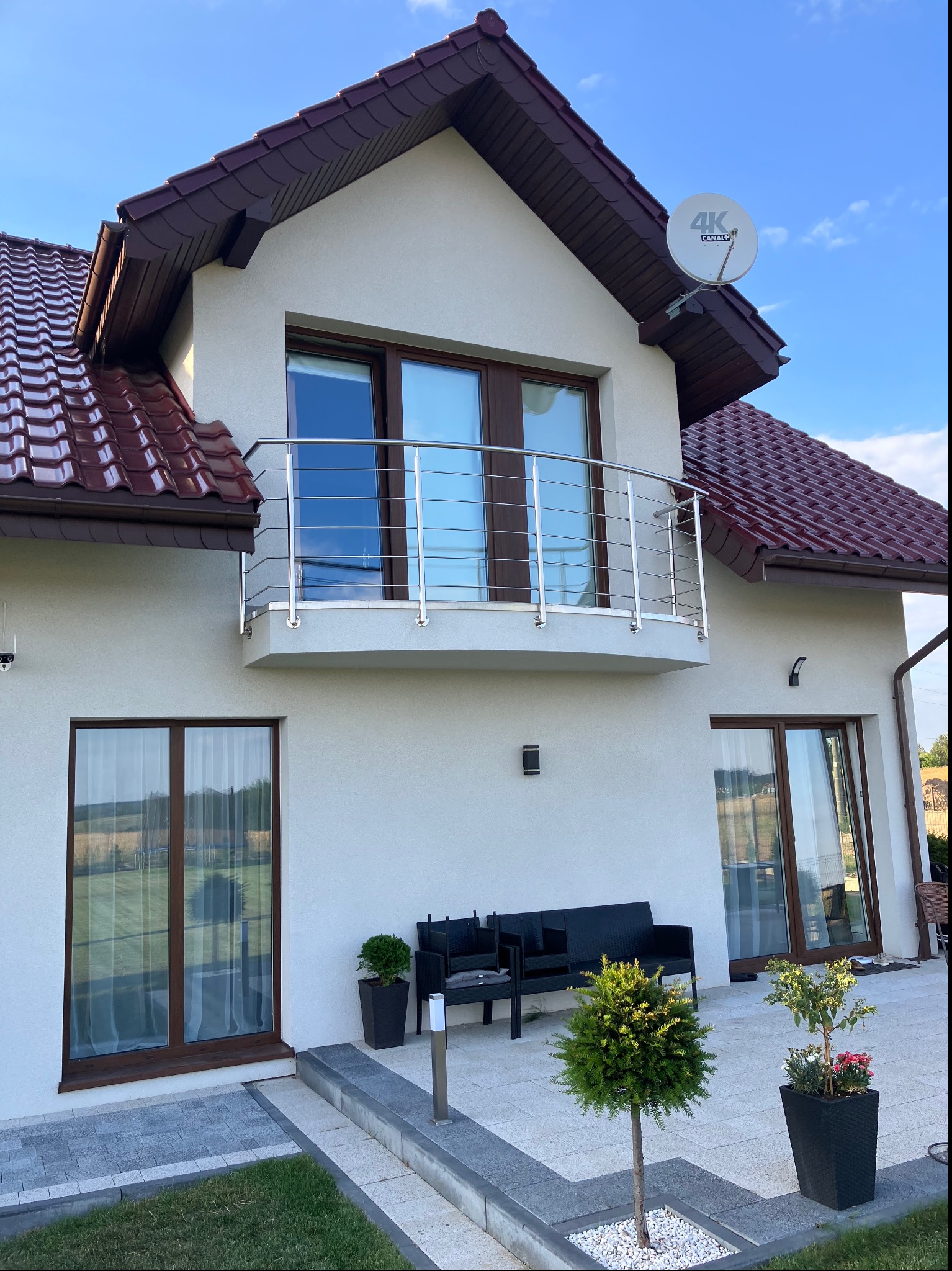 Nowoczesny dom z balkonem z balustradą ze stali nierdzewnej, brązowymi oknami i dachem z dachówki, widok z poziomu tarasu z meblami ogrodowymi i roślinami w donicach.