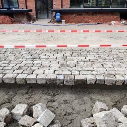 Aldis STANISŁAW GRYGORIEW - Fragment układanej nawierzchni z kostki granitowej o nieregularnych kształtach, widoczne przygotowane podłoże i zapas kostek do dalszej pracy, teren budowy zabezpieczony taśmą ostrzegawczą.