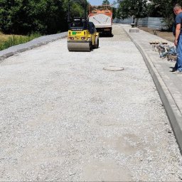 Aldis STANISŁAW GRYGORIEW - Żółty walec drogowy ubija podłoże pod kostkę na budowie chodnika, widoczna ciężarówka z piaskiem, pracownicy i ułożone fragmenty chodnika.