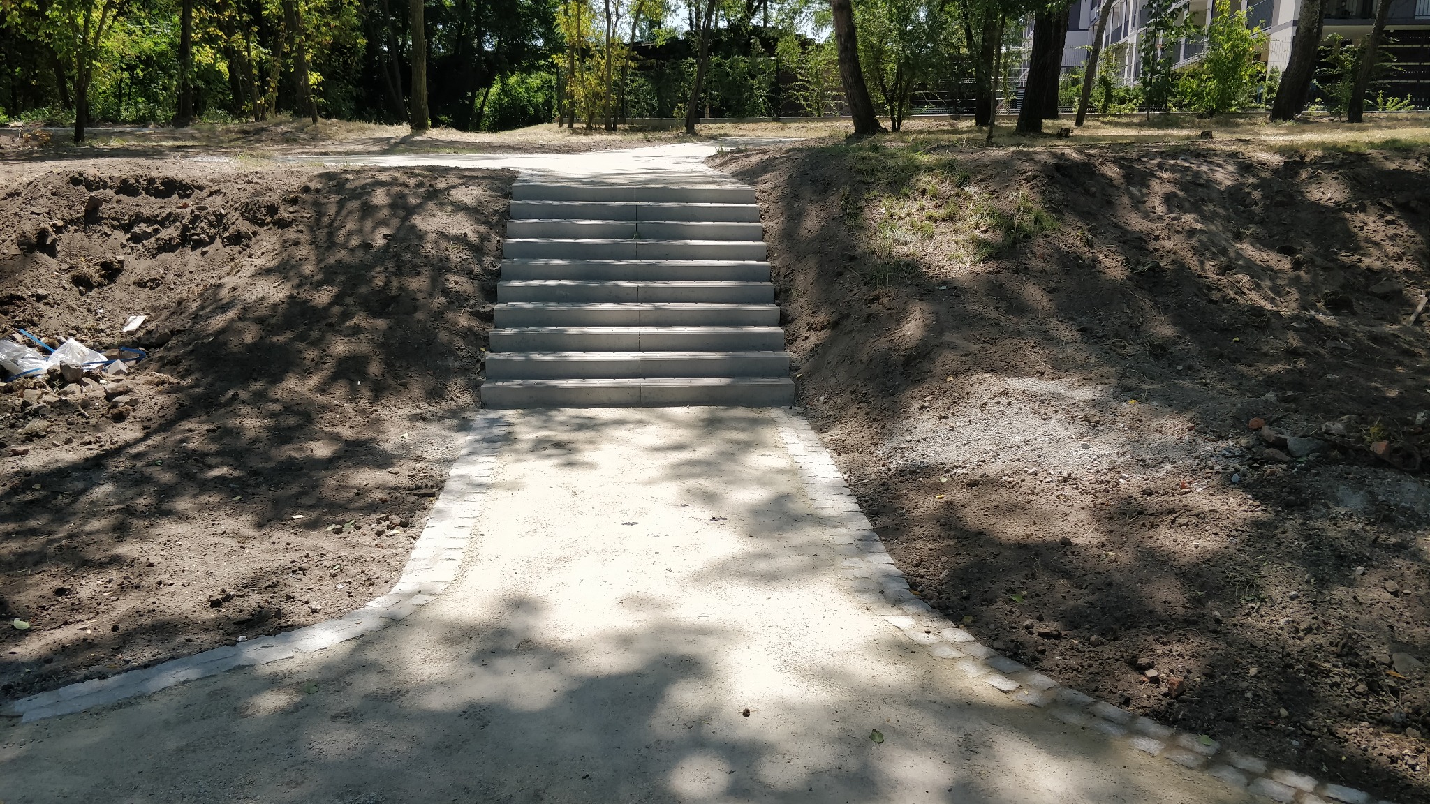 Nowo wybudowane betonowe schody prowadzące w górę zbocza, z chodnikiem z kostki po obu stronach, otoczone drzewami i ziemią.