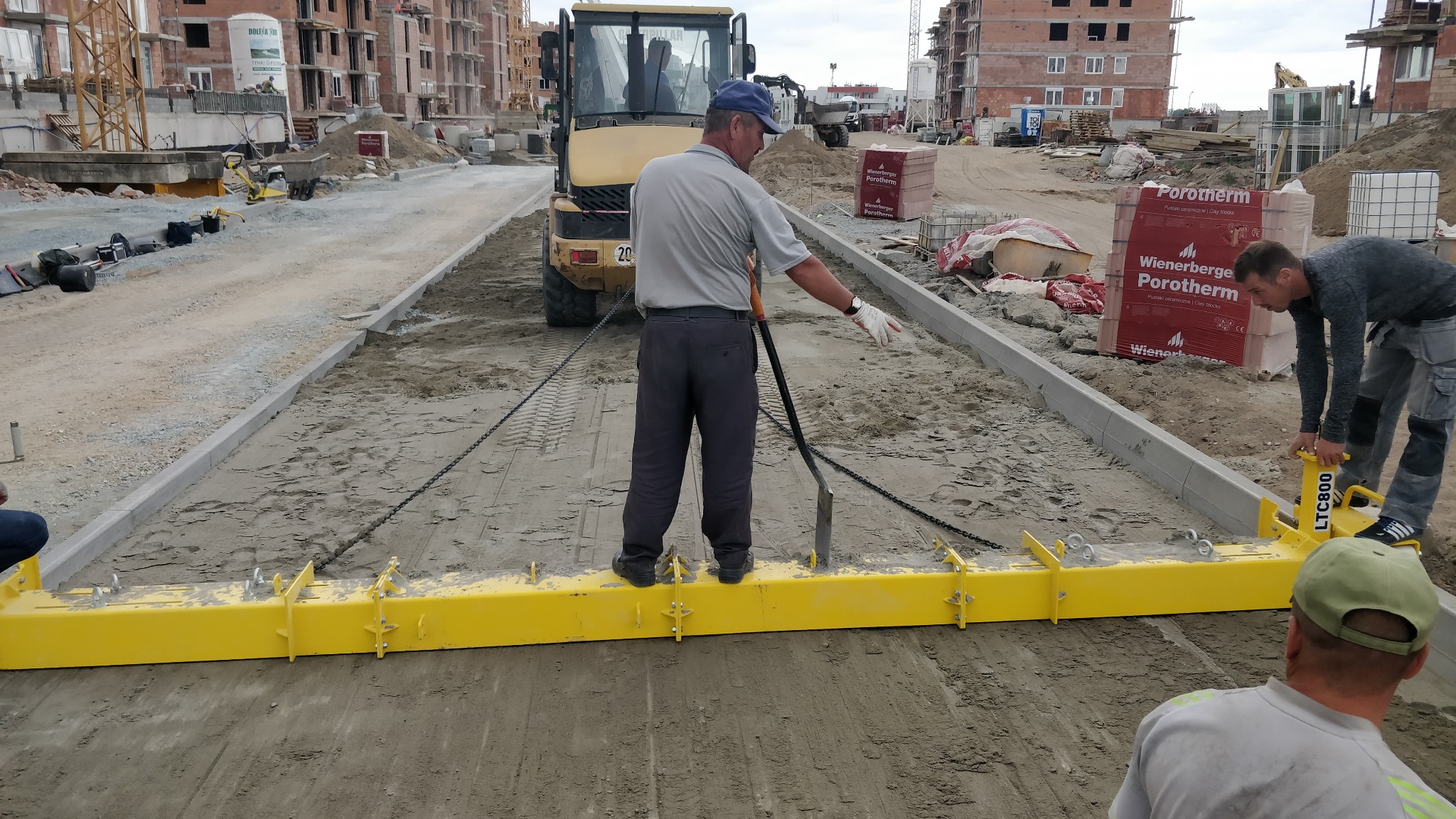 Pracownicy wyrównują podłoże pod przyszłą nawierzchnię za pomocą żółtej listwy niwelującej, w tle widać plac budowy z nowymi budynkami i maszynami budowlanymi.