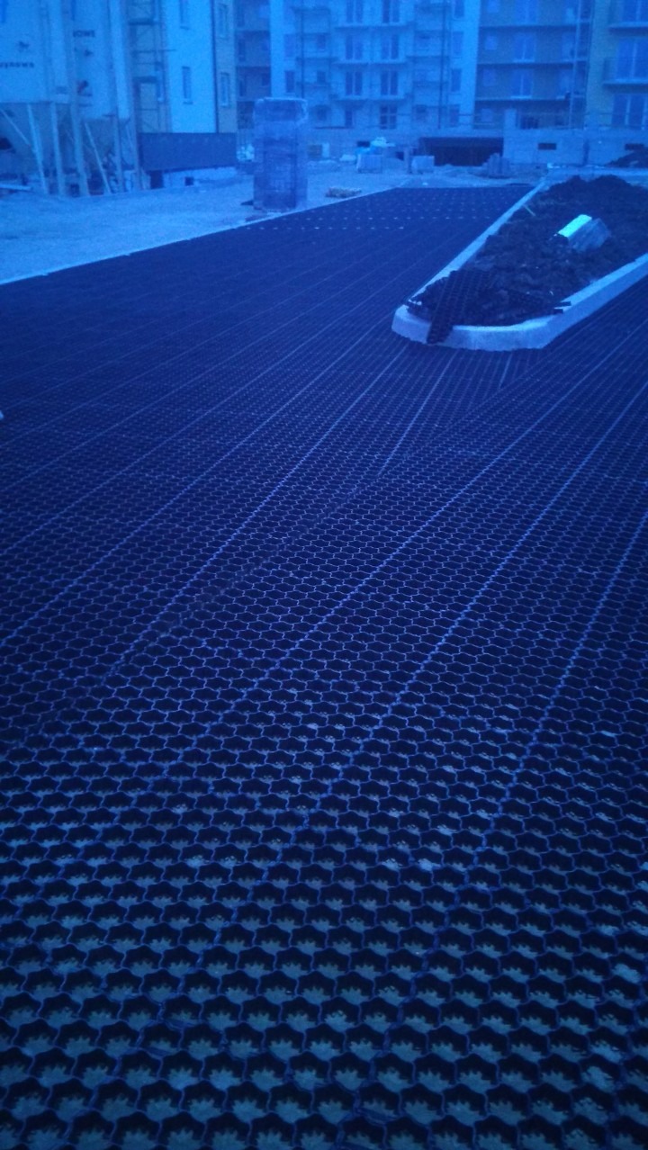 Widok z góry na ułożoną, pustą geokratę na placu budowy, z krawężnikiem i ziemią w tle, w pochmurny dzień.