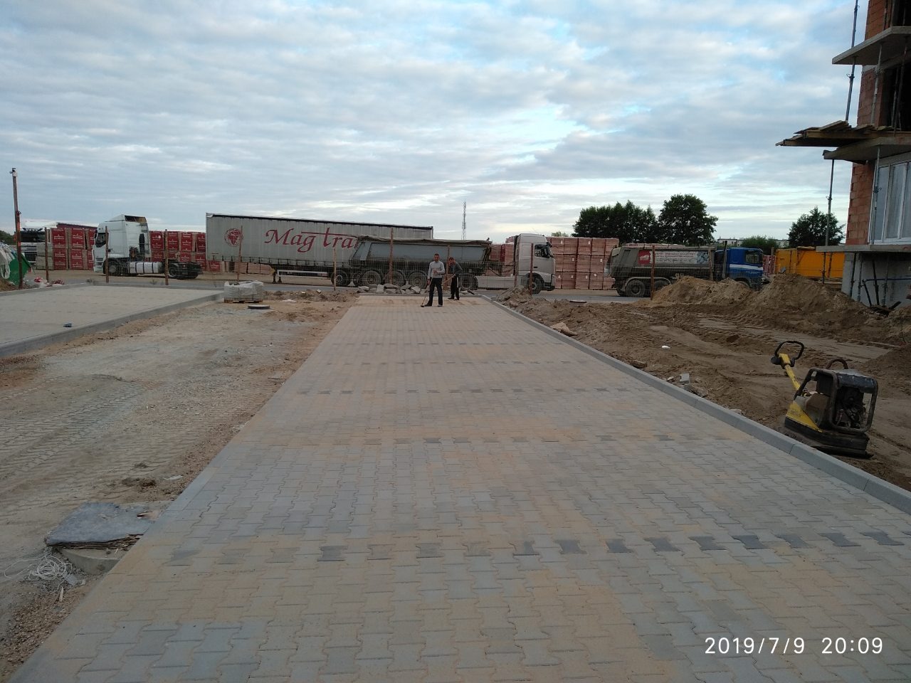 Układanie szarej kostki brukowej typu 'I', widoczny zagęszczarka wibracyjna, teren budowy z ciężarówkami w tle i pracownikami w oddali.