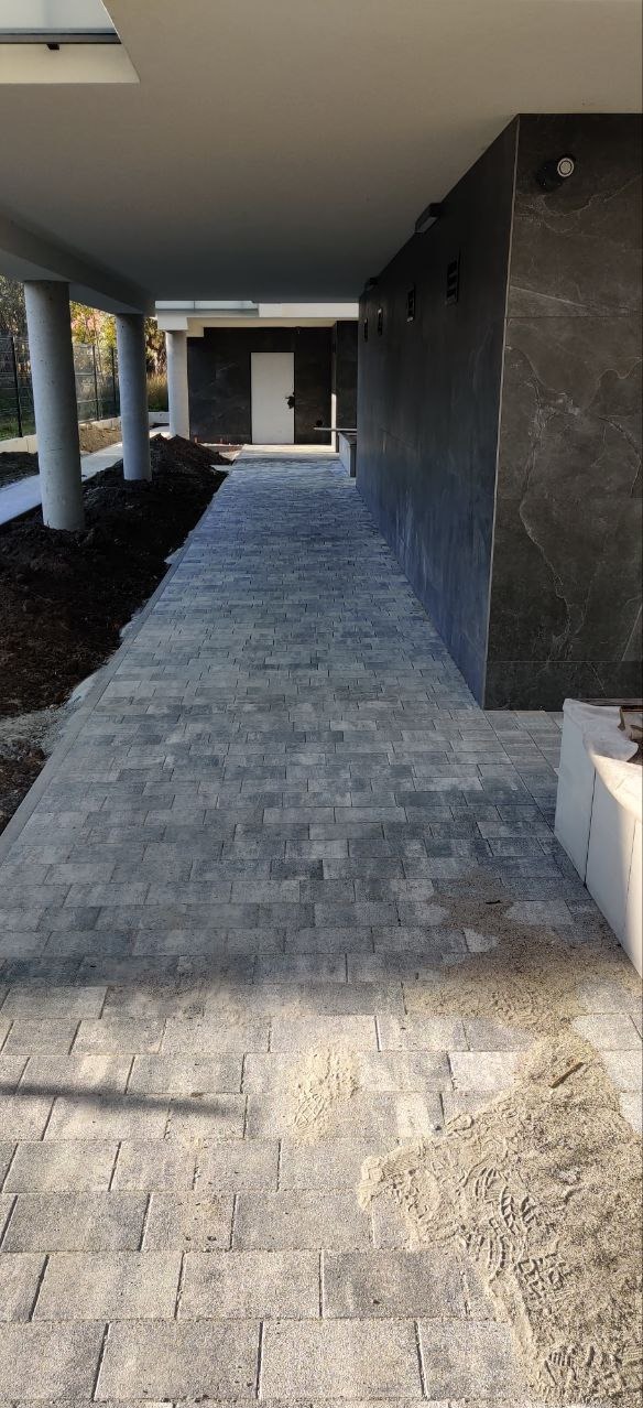 Ułożona szara kostka brukowa prowadząca do wejścia budynku z kolumnami i ciemną elewacją, widoczne ślady piasku i ziemi.