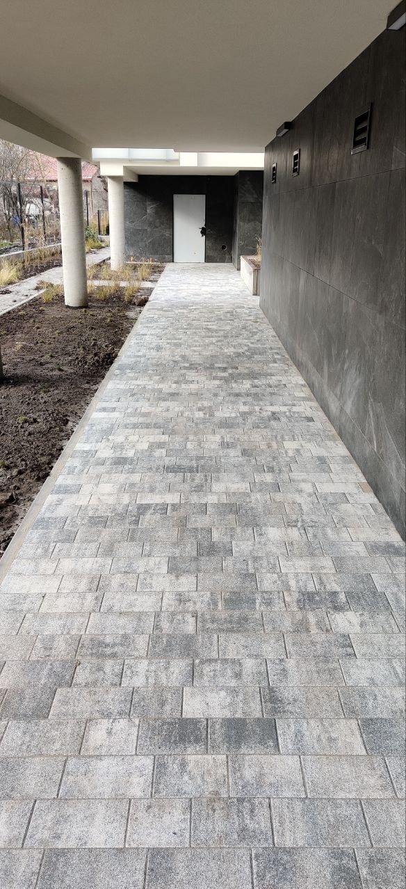 Nowoczesna ścieżka z szarej kostki brukowej o nieregularnym wzorze, prowadząca wzdłuż ciemnej, kamiennej ściany budynku z białymi kolumnami w tle.