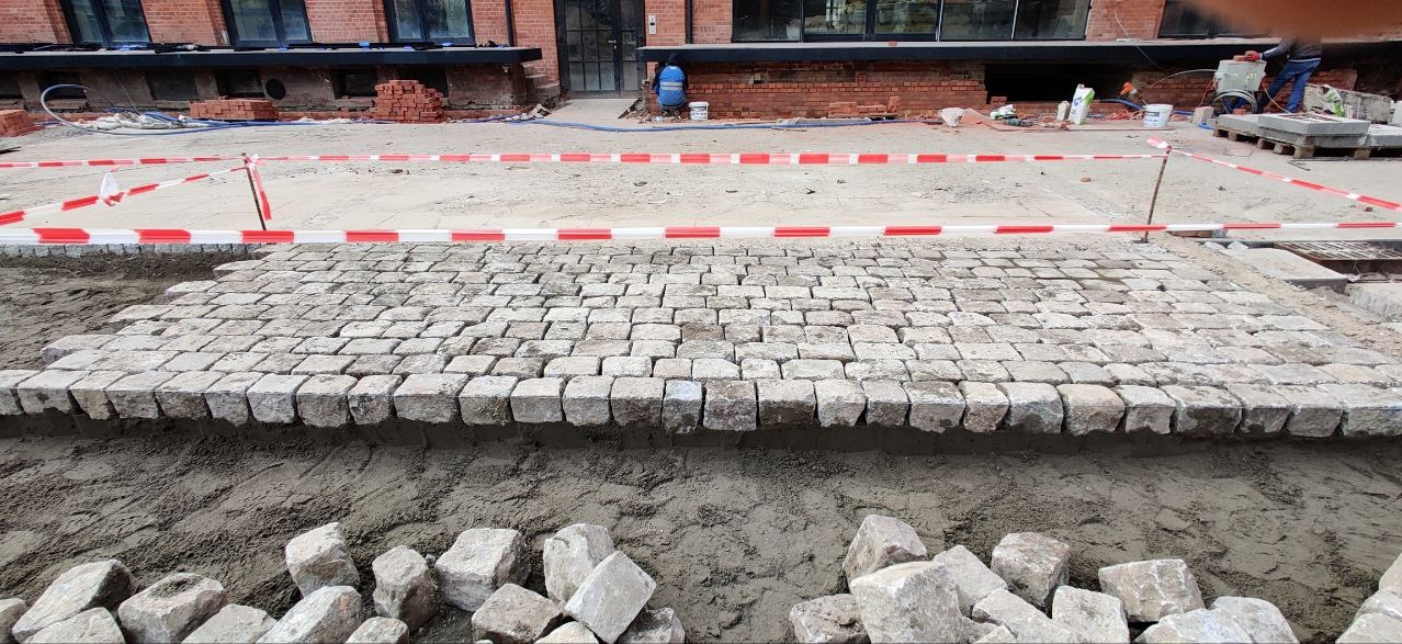 Fragment układanej nawierzchni z kostki granitowej o nieregularnych kształtach, widoczne przygotowane podłoże i zapas kostek do dalszej pracy, teren budowy zabezpieczony taśmą ostrzegawczą.