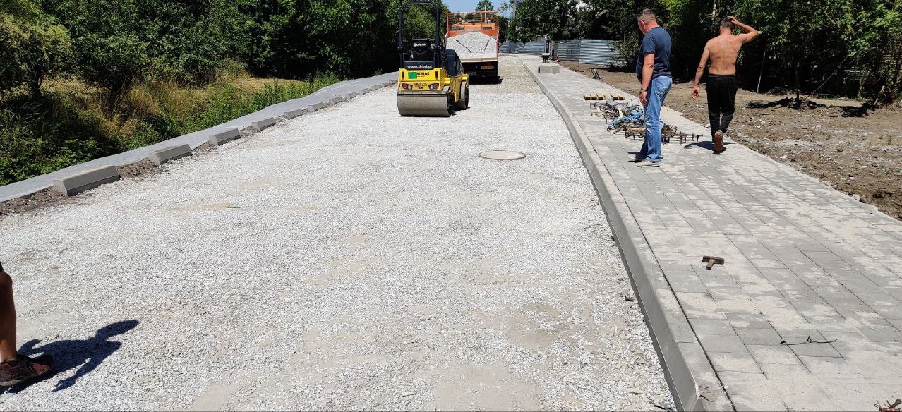 Żółty walec drogowy ubija podłoże pod kostkę na budowie chodnika, widoczna ciężarówka z piaskiem, pracownicy i ułożone fragmenty chodnika.