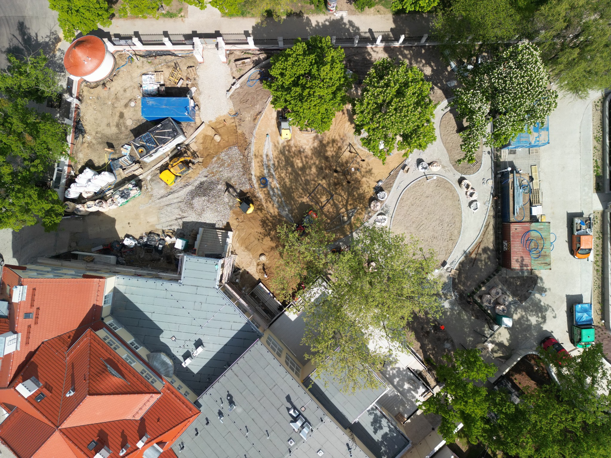 Widok z góry na plac budowy z żółtą koparką, teren przygotowany pod kostkę brukową, ułożone ścieżki i piaskownica. Częściowo widoczne budynki z czerwoną dachówką.