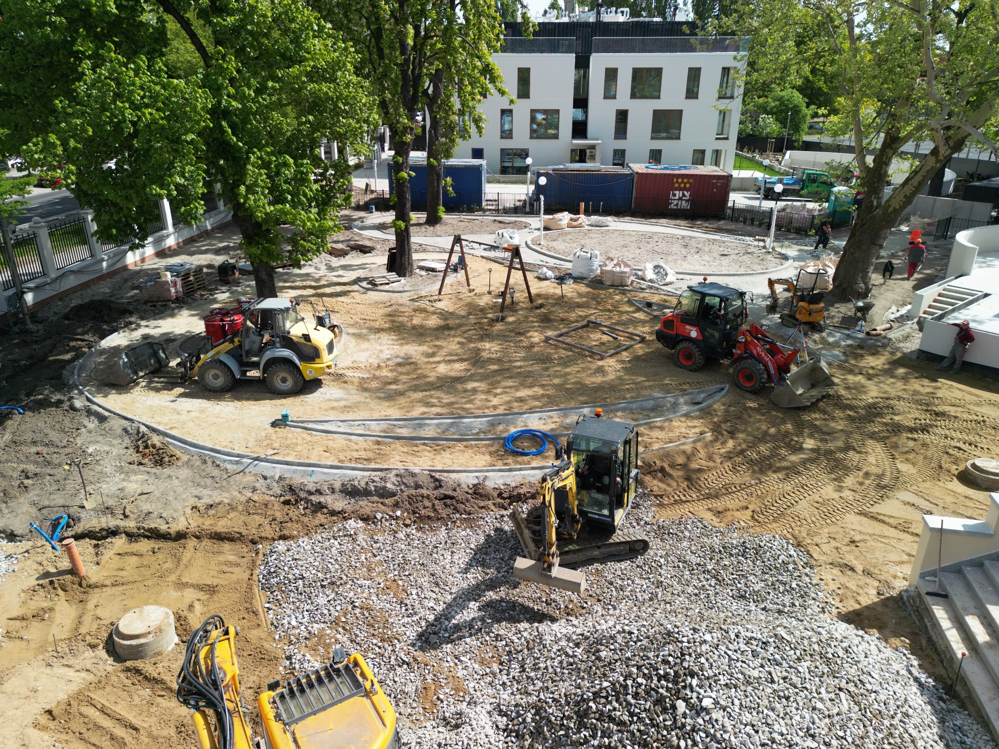 Plac budowy z maszynami: koparka, ładowarka. Teren przygotowany pod kostkę brukową, widoczne obrzeża. W tle plac zabaw i budynek.