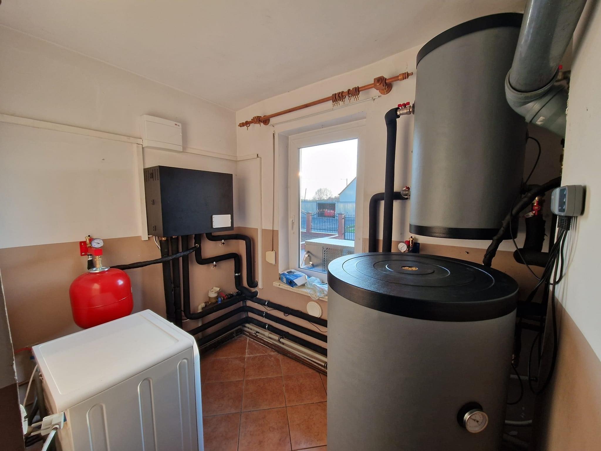 Kotłownia z szarym zasobnikiem ciepłej wody, czarnym kotłem grzewczym, czerwonym zbiornikiem wyrównawczym, białą pralką i rurami grzewczymi na brązowych płytkach podłogowych.