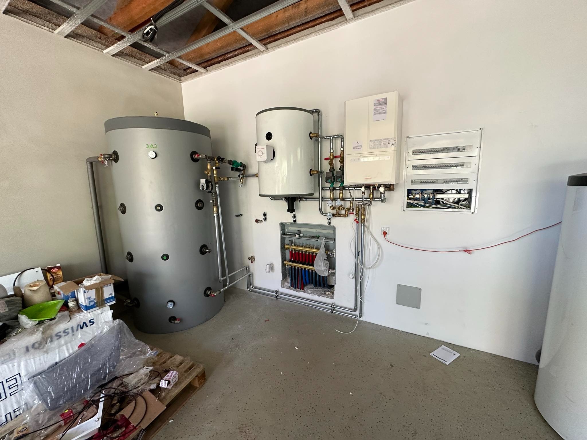 Szara akumulacyjna butla ciepłej wody użytkowej oraz biały zasobnik ciepłej wody, połączone rurami z rozdzielaczem w ścianie i kotłem gazowym, widoczne również skrzynki elektryczne, surowy stan...