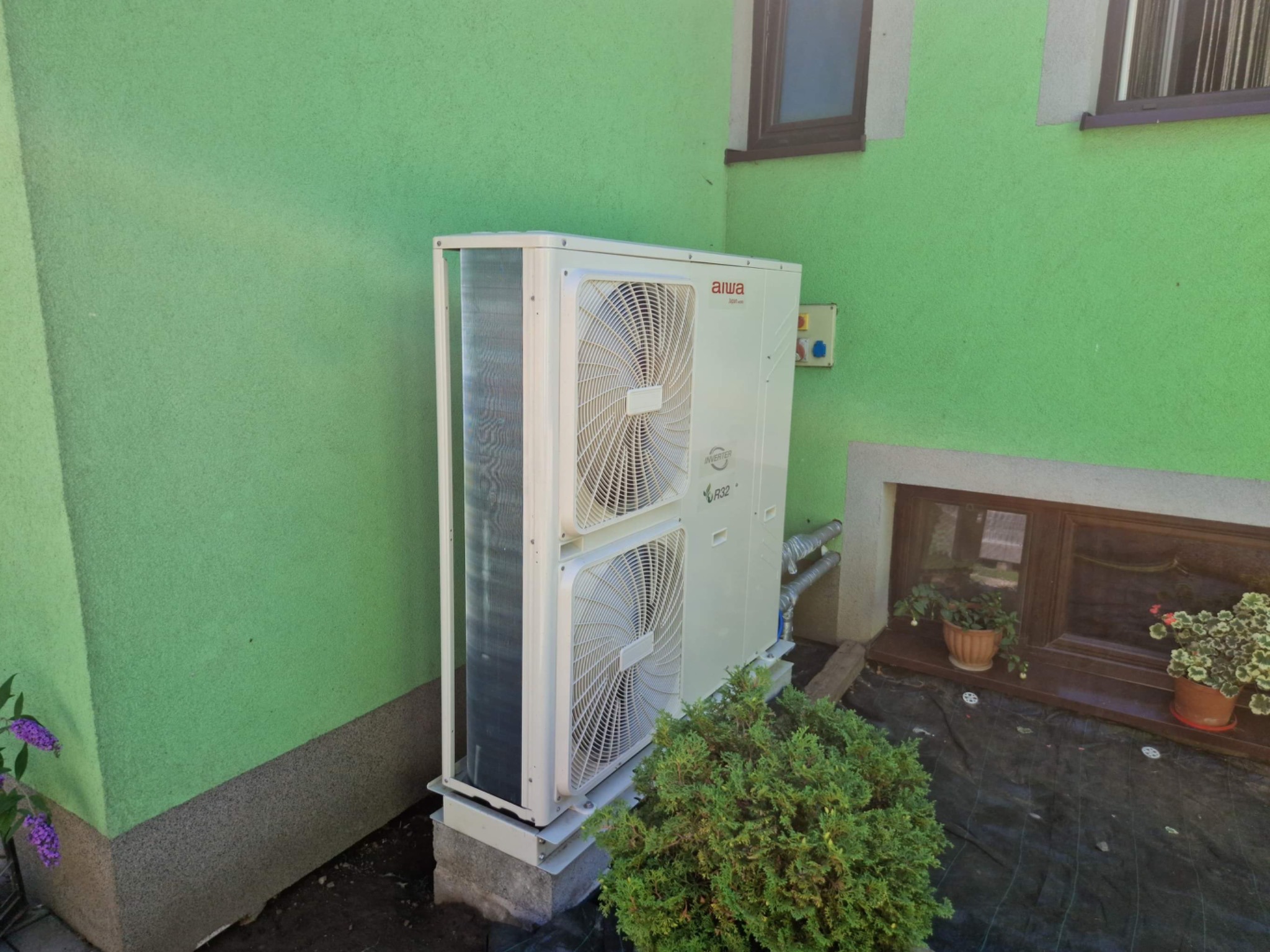 Biała jednostka zewnętrzna klimatyzacji marki Aiwa, zamontowana przy zielonej ścianie domu, z widocznymi rurami i krzewem z przodu.