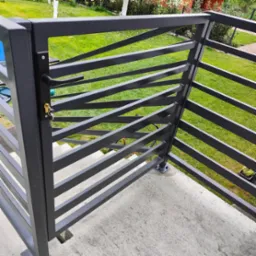 Szara metalowa furtka z ukośnymi szczeblami, zamykana na klucz, prowadząca na schody, widok z góry na trawnik.