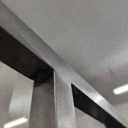 Szczegół spawu na konstrukcji z profili metalowych, widoczne ślady szlifowania, surowy metal w szarym odcieniu, oświetlenie warsztatowe w tle.