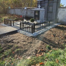 Artystal Paweł Krzan - Czarne, metalowe ogrodzenie wokół nagrobka, wykończone drobnym grysem, z brukowanym chodnikiem i drabiną opartą o mur w tle.