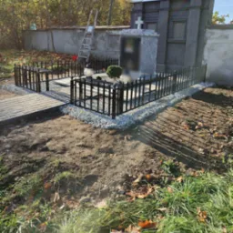 Czarne, metalowe ogrodzenie wokół nagrobka, wykończone drobnym grysem, z brukowanym chodnikiem i drabiną opartą o mur w tle.