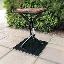 Artystal Paweł Krzan - Stolik kawowy z drewnianym blatem i geometryczną podstawą z czarnego metalu, ustawiony na brukowanej powierzchni przed zielonym żywopłotem.