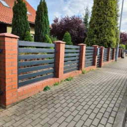 Nowoczesne ogrodzenie metalowe z poziomymi panelami w kolorze grafitowym, osadzone na ceglanych słupkach, widok z perspektywy chodnika z kostki brukowej, w tle dom z czerwoną dachówką i zielone...