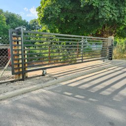 Artystal Paweł Krzan - Suwana brama wjazdowa z poziomych metalowych profili, zamontowana na metalowych słupkach, z widocznymi kółkami jezdnymi na tle zieleni i asfaltowej drogi.