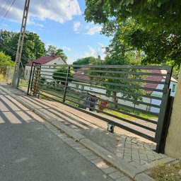 Artystal Paweł Krzan - Przesuwna brama wjazdowa z poziomych profili, zamontowana na kółkach, widok z zewnątrz posesji, w tle dom z czerwonym dachem i drzewa.