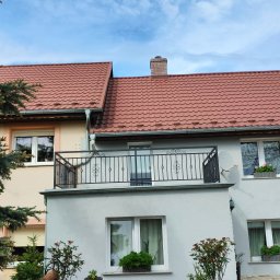 Artystal Paweł Krzan - Metalowa balustrada balkonowa z ozdobnymi elementami na tle błękitnego nieba, widok na dom z czerwonym dachem i roślinami w donicach.