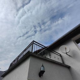 Artystal Paweł Krzan - Czarna balustrada balkonowa z metalowymi zdobieniami, zamontowana na białym budynku z widoczną lampą zewnętrzną i anteną satelitarną na tle pochmurnego nieba.