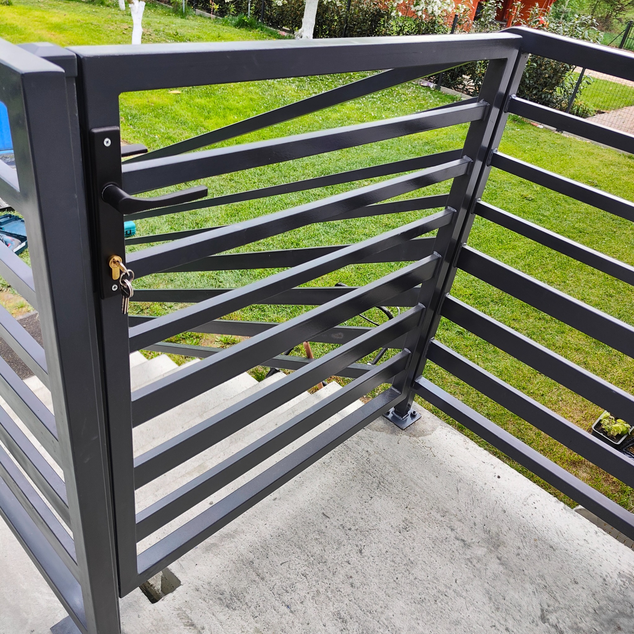 Szara metalowa furtka z ukośnymi szczeblami, zamykana na klucz, prowadząca na schody, widok z góry na trawnik.