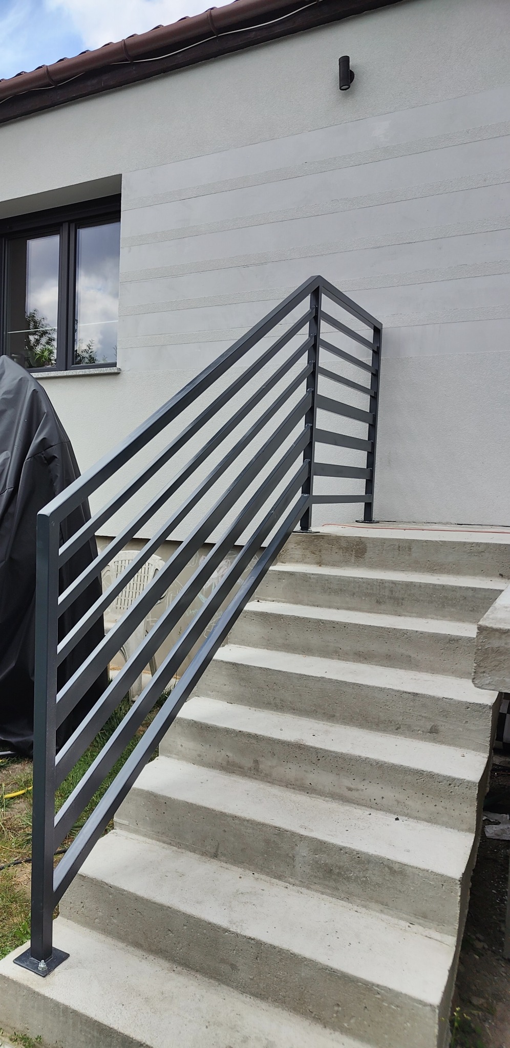 Zewnętrzne schody betonowe z nowoczesną metalową poręczą z poziomych profili, widok z dołu na tle ściany budynku z oknem.